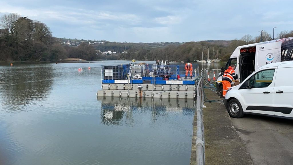 Drilling rig set up on a pontoon on the River Teifi, with vans on the path to the right of the river.