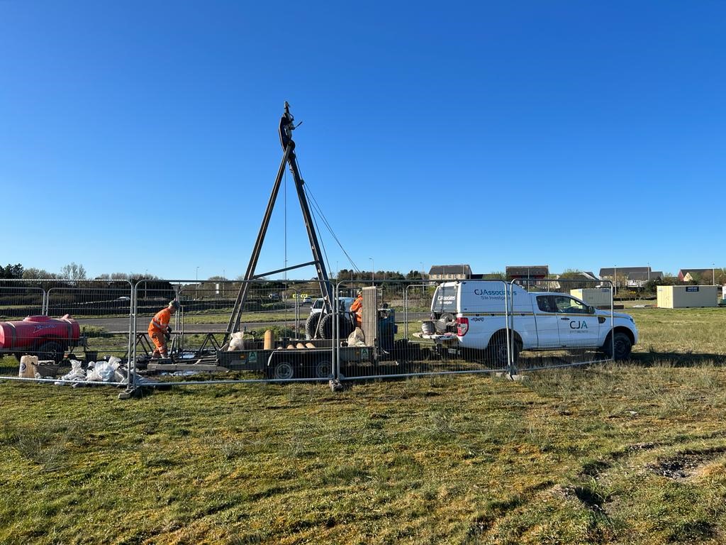 Cable percussion rig set up in a field at Llanelli Wellness and Life Science village, with workers in hi vis PPE operating the rig, and a CJ Associates hilux vehicle on site.