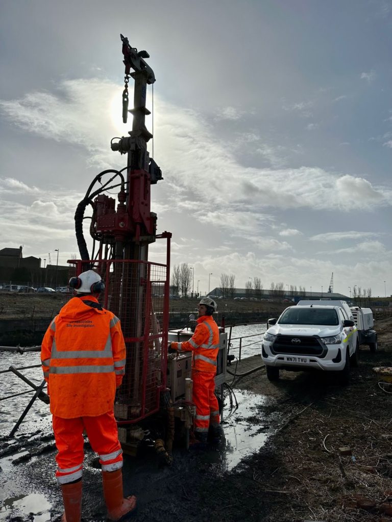 Two workers dressed in hi-vis PPE, operating a drilling rig, with a hilux vehicle in the background and the sky.