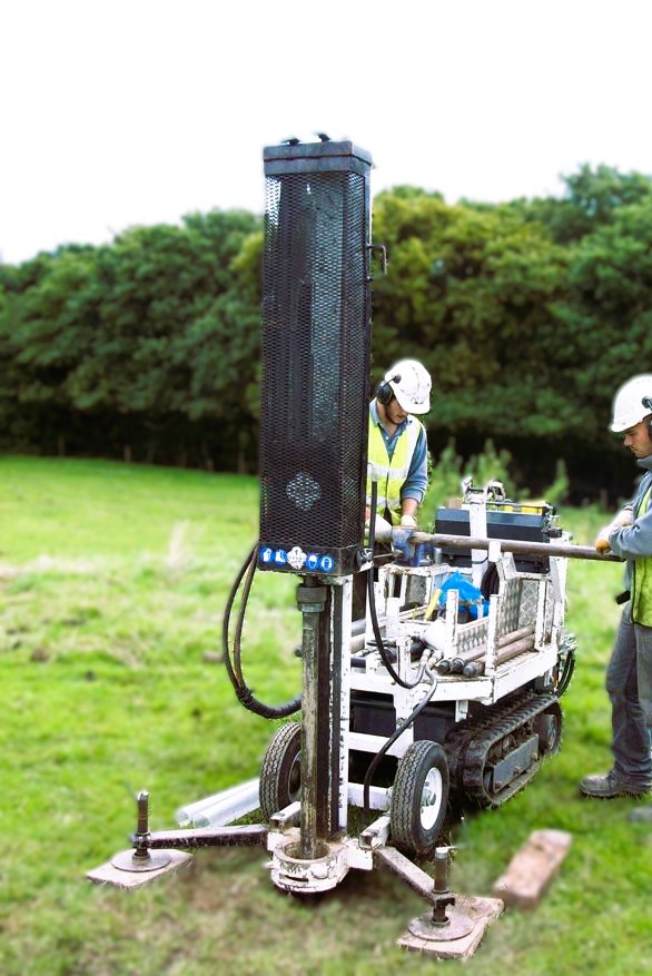 Two CJ Associates site workers operating a windowless sampling rig, in a field, dressed in hi-vis and personal protective clothing and equipment.