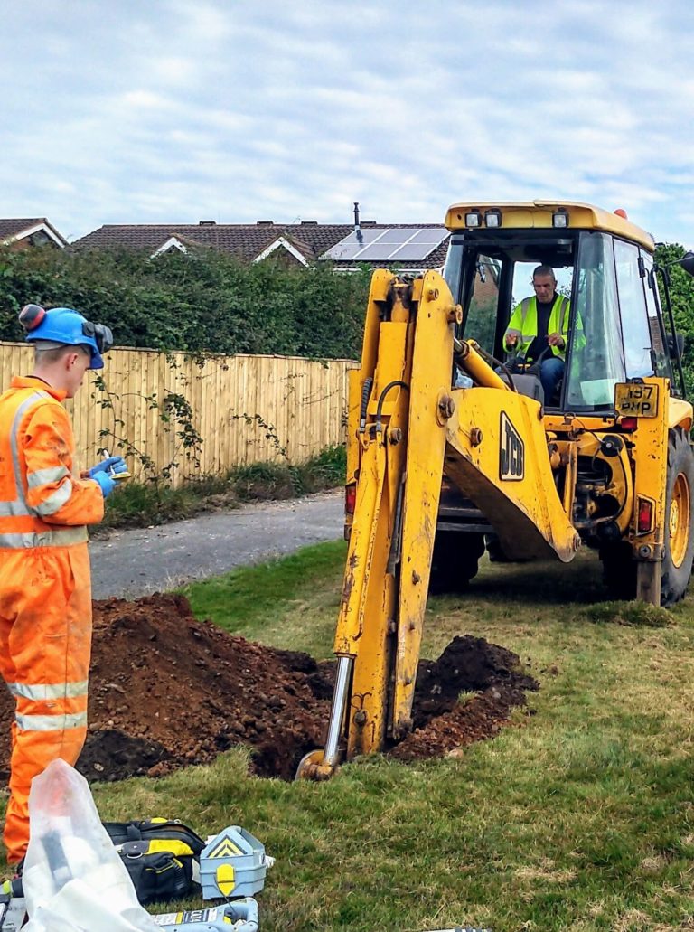 A worker dressed in hi-vis personal protective clothing, operating a mechanical excavator, digging a trial pit, with another worker dressed in hi-vis personal protective clothing stood opposite from the JCB.