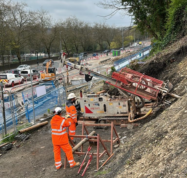 Workers dressed in hi-vis personal protective clothing, operating a CJ Associates drilling rig on a slope, with the roadside and vehicles in the background.