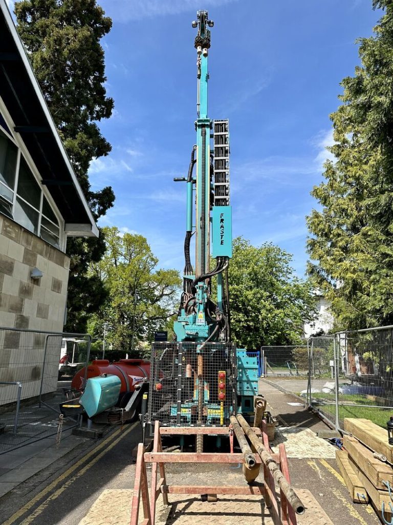 A CJ Associates rotary drilling rig, set out in the middle of a concrete road, with the blue sky and trees in the background.