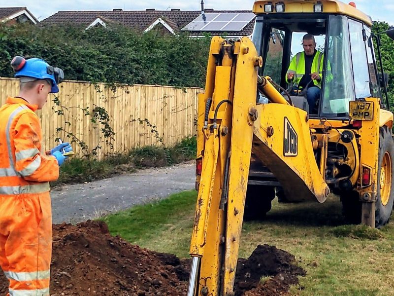 A worker dressed in hi-vis personal protective clothing, operating a mechanical excavator, digging a trial pit, with another worker dressed in hi-vis personal protective clothing stood opposite from the JCB.